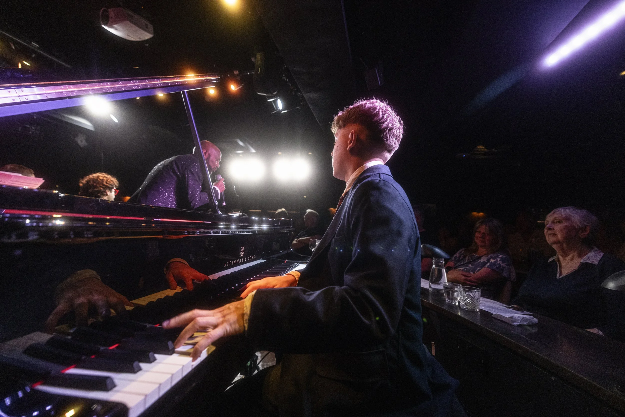 A young man in a dark suit playing a grand piano in a dimly lit room with a performer singing into a microphone. Audience members are seated nearby, some smiling, with drinks on the table.