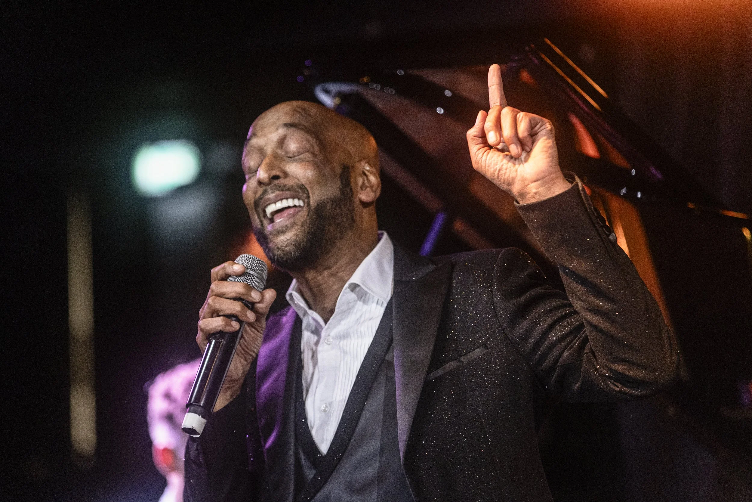 Tommy Blaize passionately singing into a microphone with his eyes closed and one finger raised, illuminated by warm stage lighting.