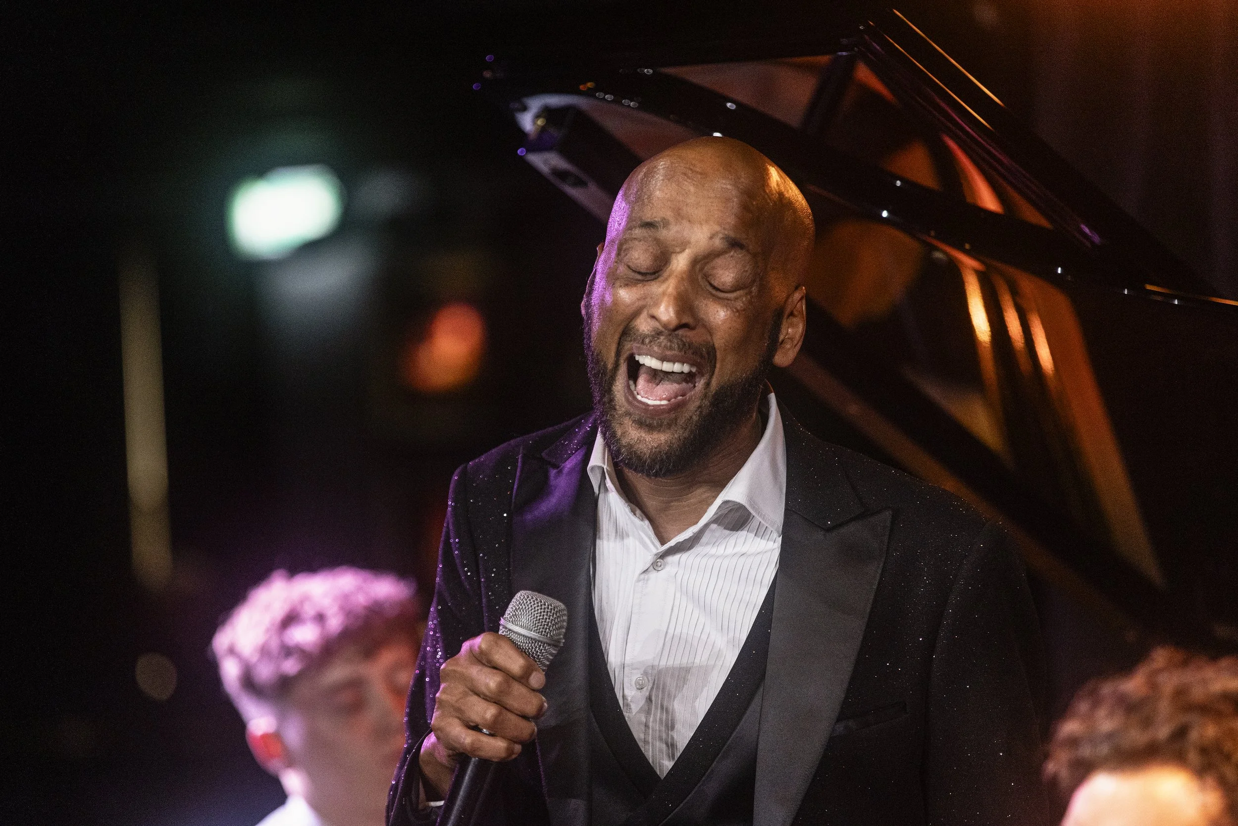 Tommy Blaize singing passionately into a microphone at a concert or event, with a grand piano in the background and people in the foreground.