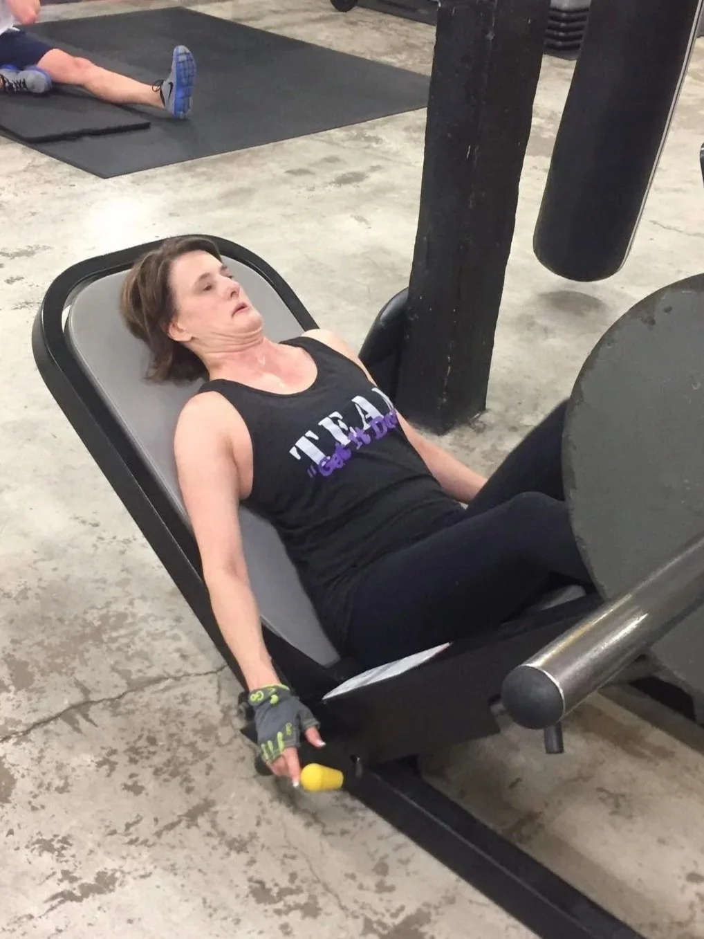 A woman lying on a workout bench at a gym, after an intense workout, with a fatigued expression, holding a yellow grip strengthener in her right hand.