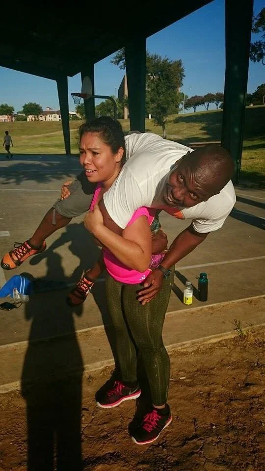 A woman is holding a young girl on her back at an outdoor basketball court. The woman is smiling, wearing athletic clothing, and the girl appears to be comfortable. The background shows a park with trees and buildings.