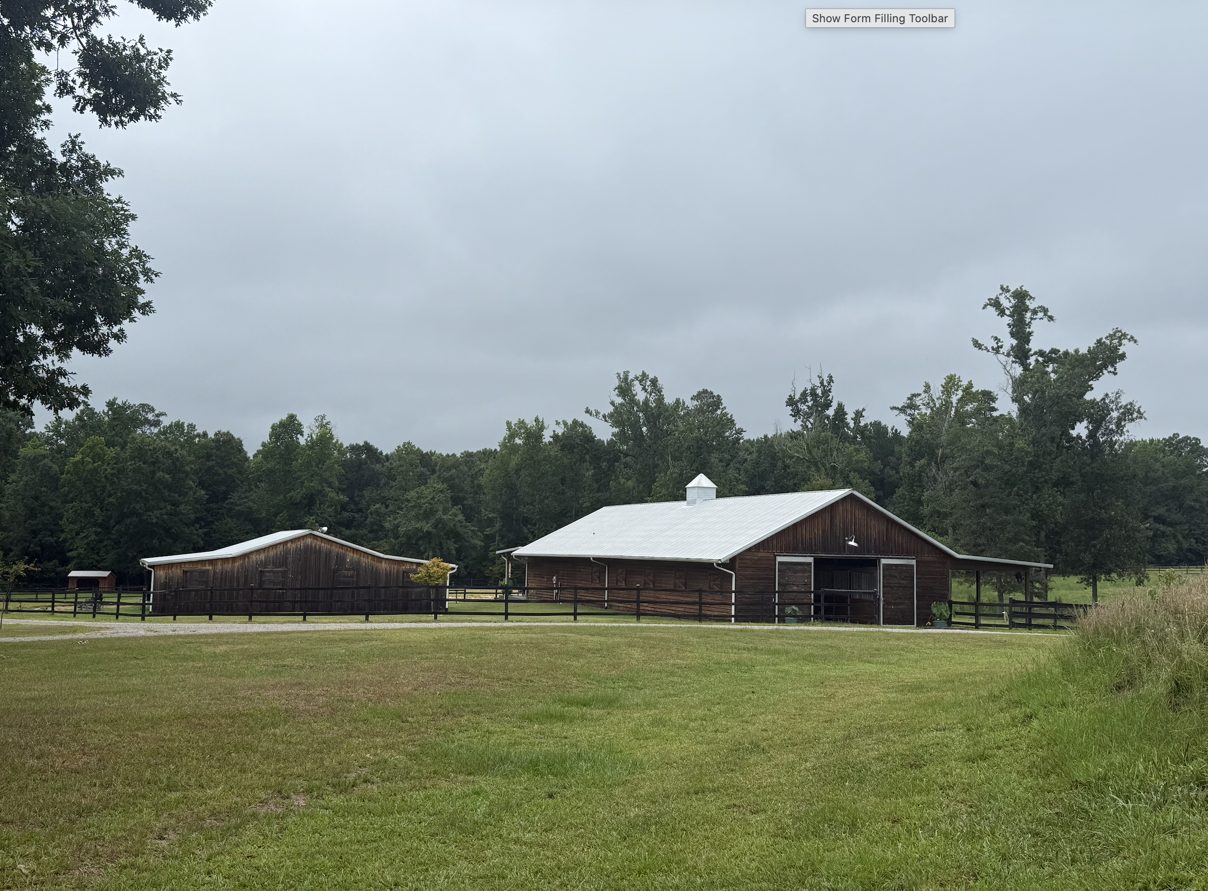 Overcast sky above a rural farm with two brown wooden barns, surrounded by green trees and a grassy field.