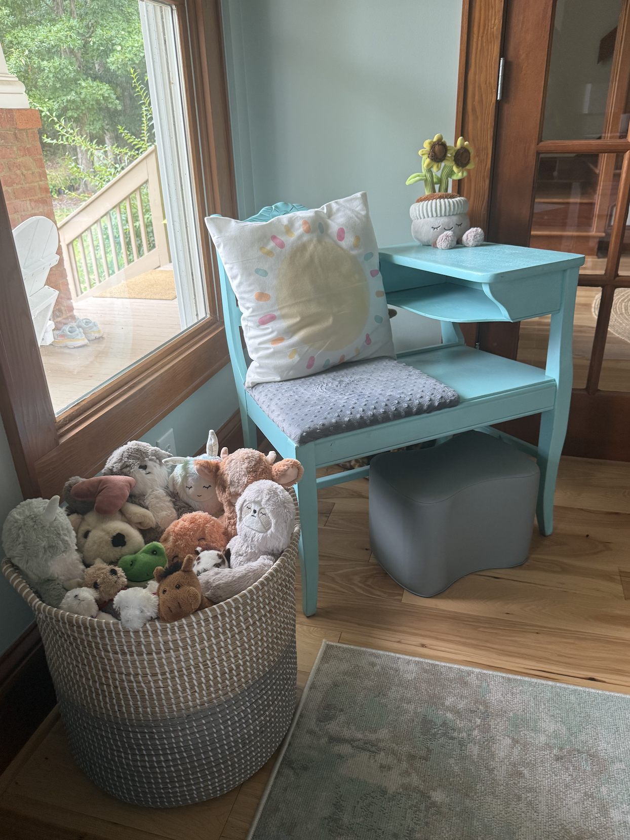 A cozy corner featuring a light blue wooden bench with a gray plush cushion and a decorative pillow with a sun design. A small turquoise side table with a plush flowerpot with sunflowers sits beside the bench. A basket filled with various stuffed animals, including a dog, llama, unicorn, frog, and others, is positioned next to the bench. The scene is next to a large window with a view of a porch and greenery outside.