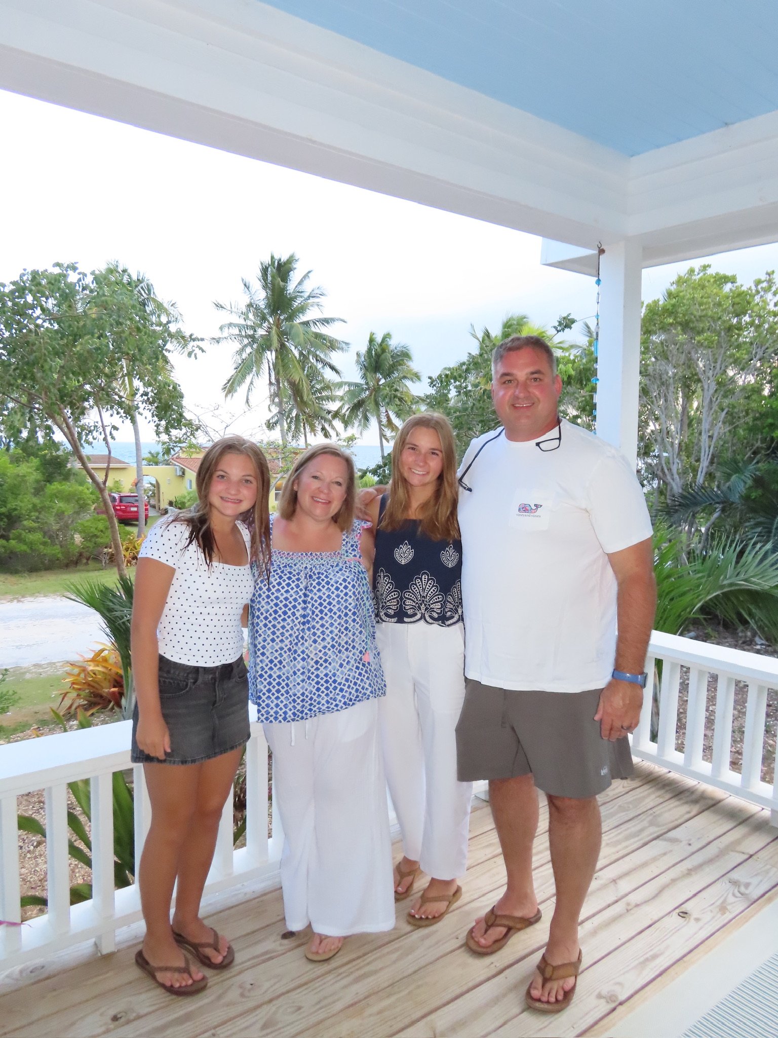 A group of five people, three females and one male, smiling and standing together on a porch with a tropical background, including palm trees and green foliage.