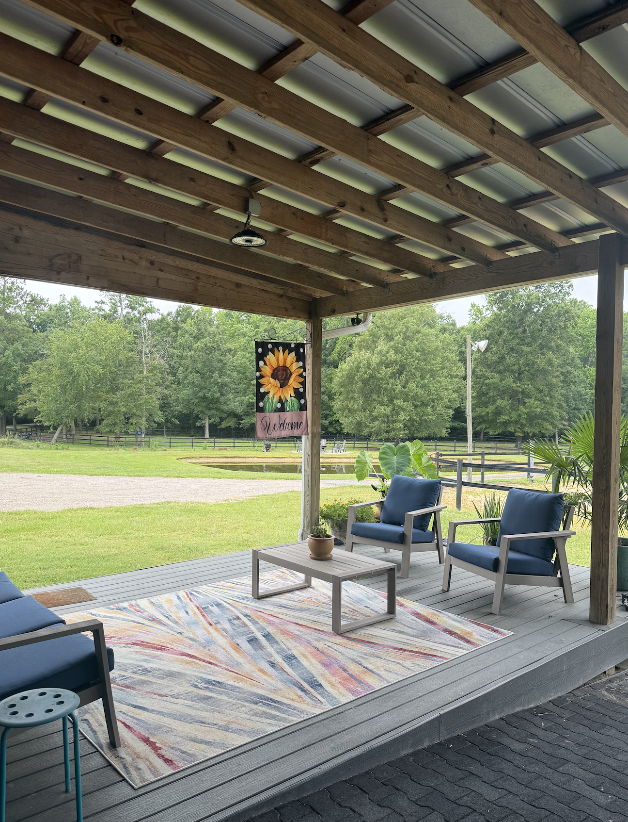 A covered outdoor patio with blue cushioned chairs, a coffee table, a colorful rug, and potted plants. In the background, green trees and a grassy area are visible, along with a sign that says 'Welcome' with a sunflower design.