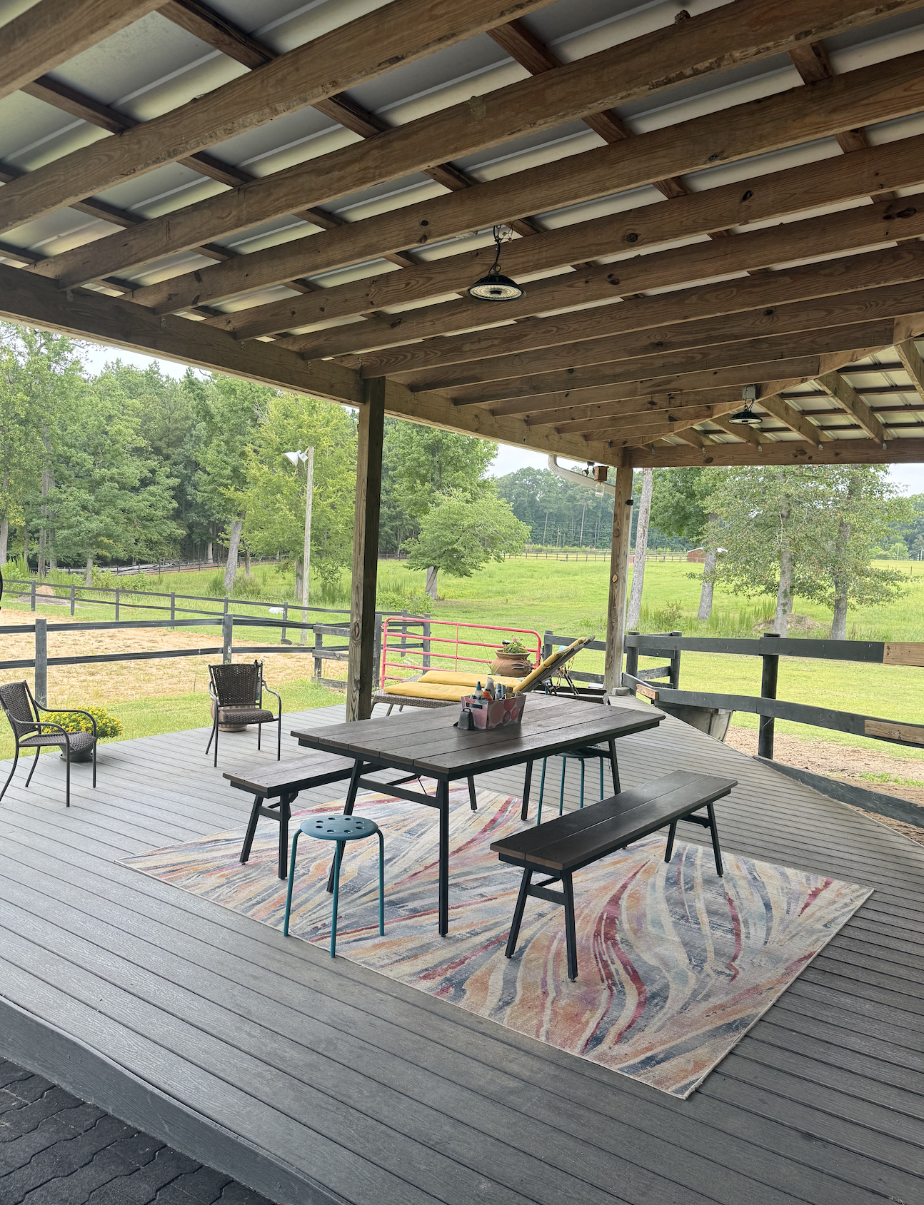 Covered outdoor deck with a wooden table, chairs, and a colorful rug, overlooking a green field with trees and fencing.