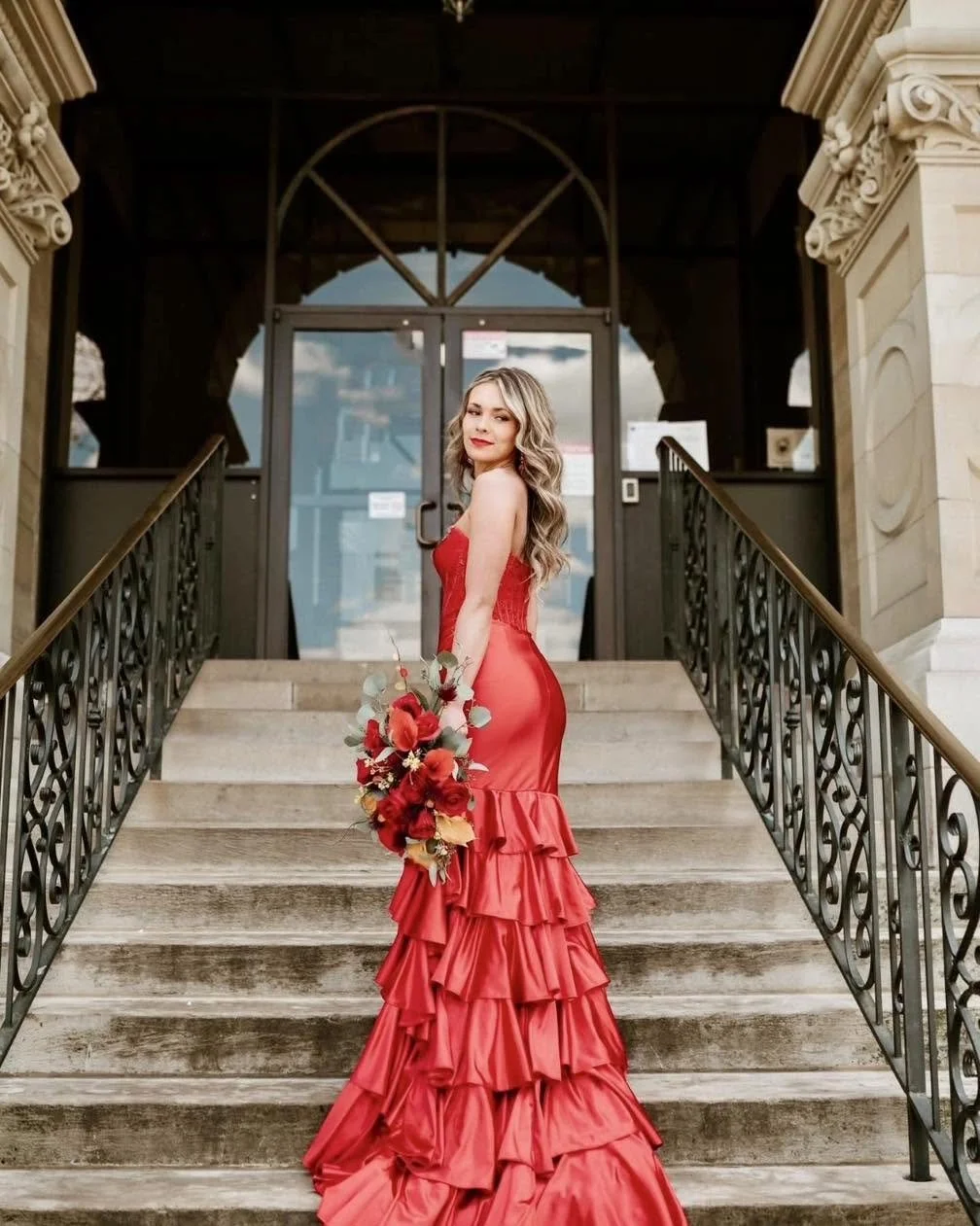 A woman in a red ruffled gown holding a red floral bouquet stands on a stone staircase outside a building with ornate architectural details.