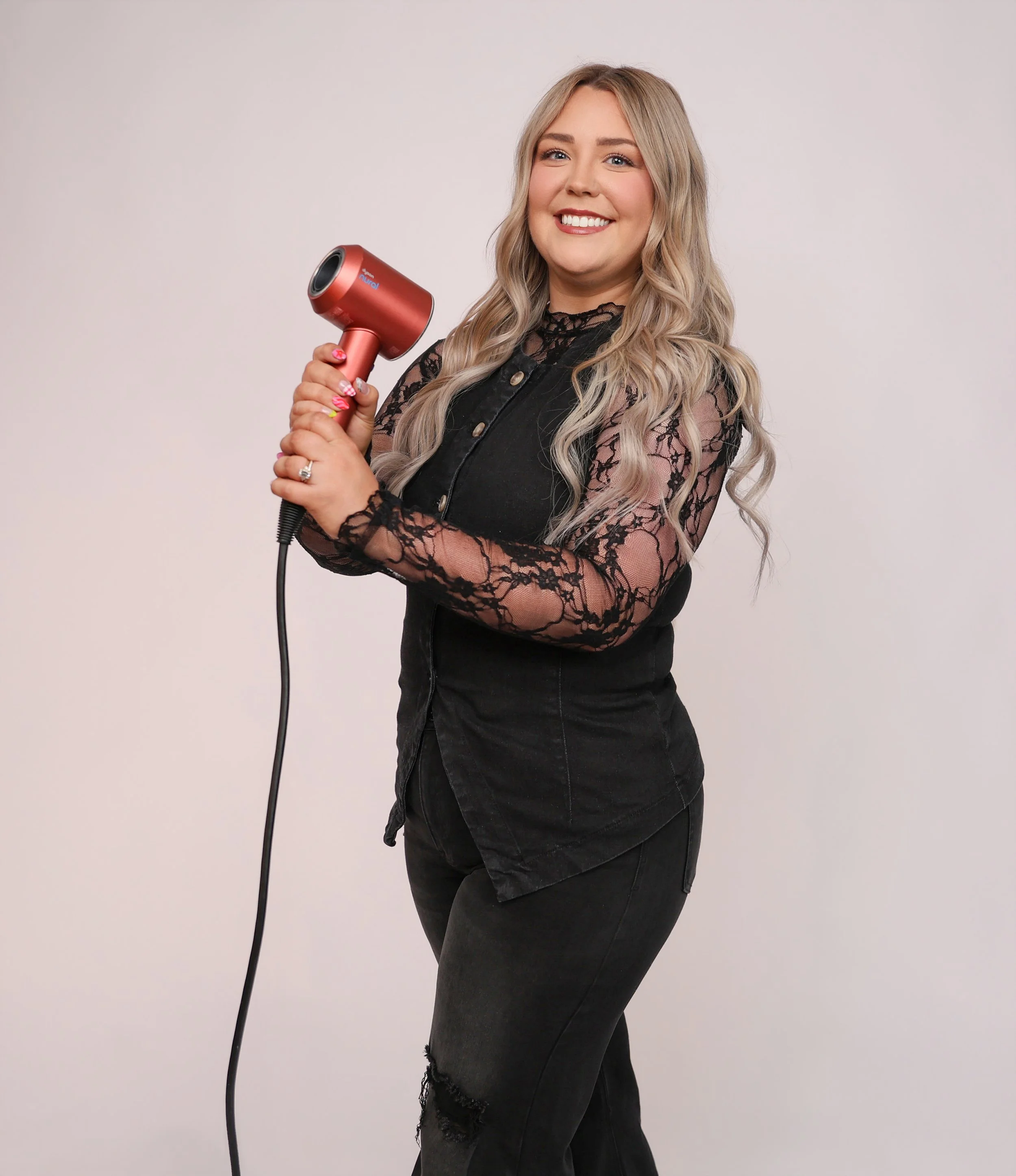 A smiling woman with long wavy blonde hair holding a red hairdryer, wearing a black lace top and black distressed jeans, standing against a plain light gray background.