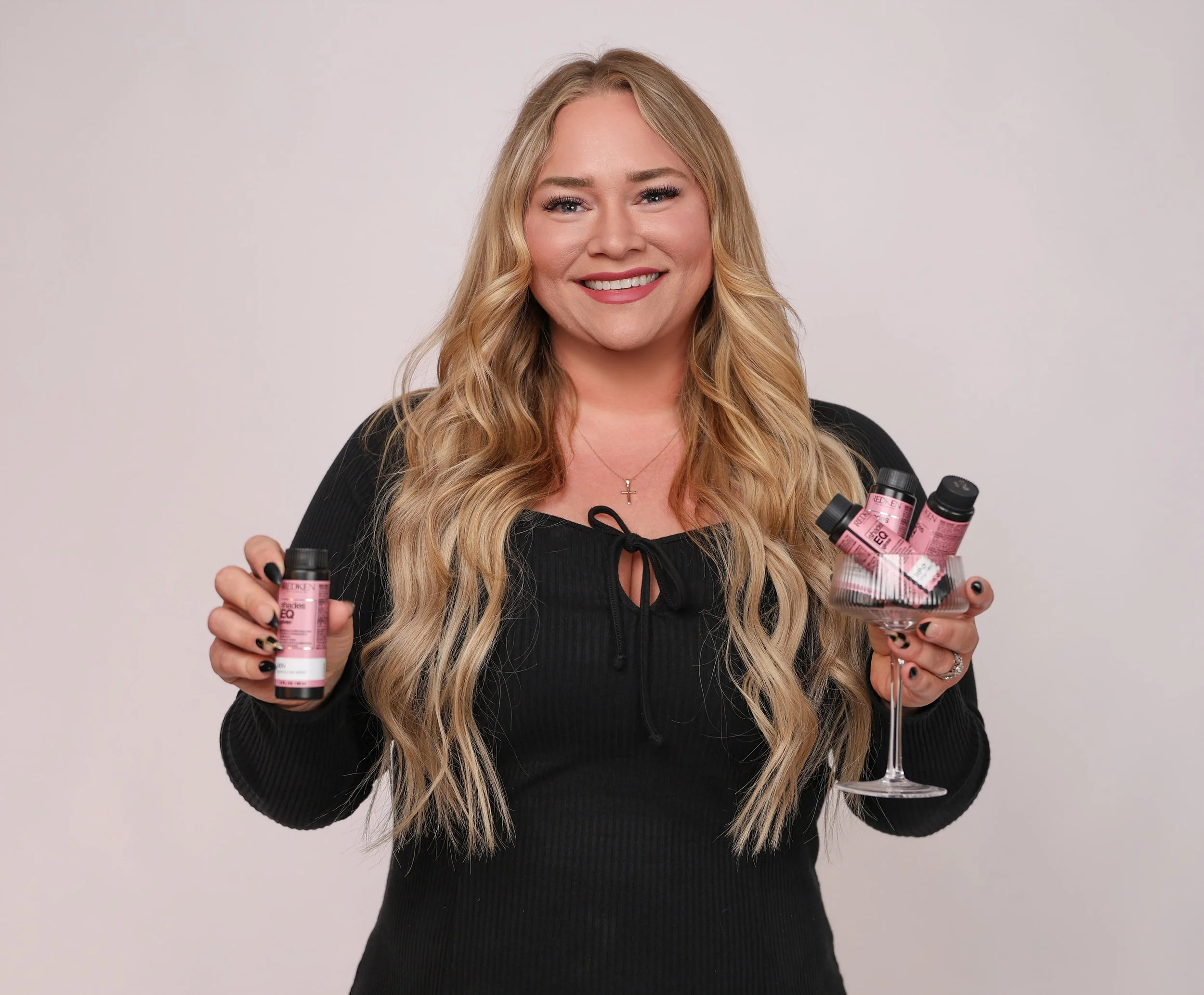 Woman with long blonde hair, smiling, holding bottles of pink hair dye in one hand and standing next to a wine glass filled with hair dye bottles.