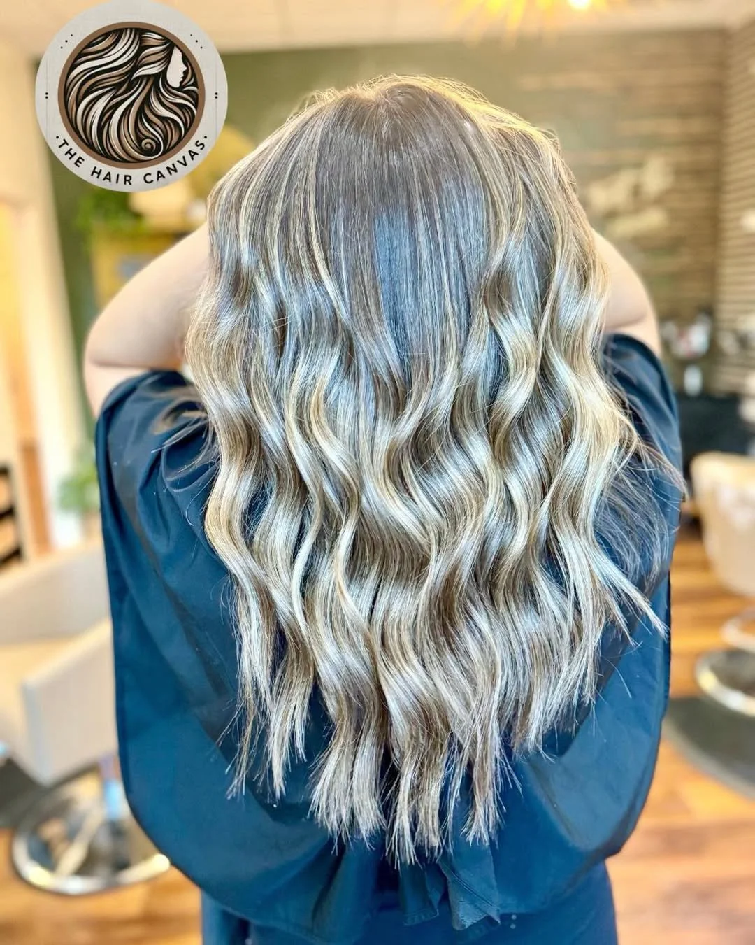Long wavy blonde hair with darker roots and highlights view from the back in a salon setting.