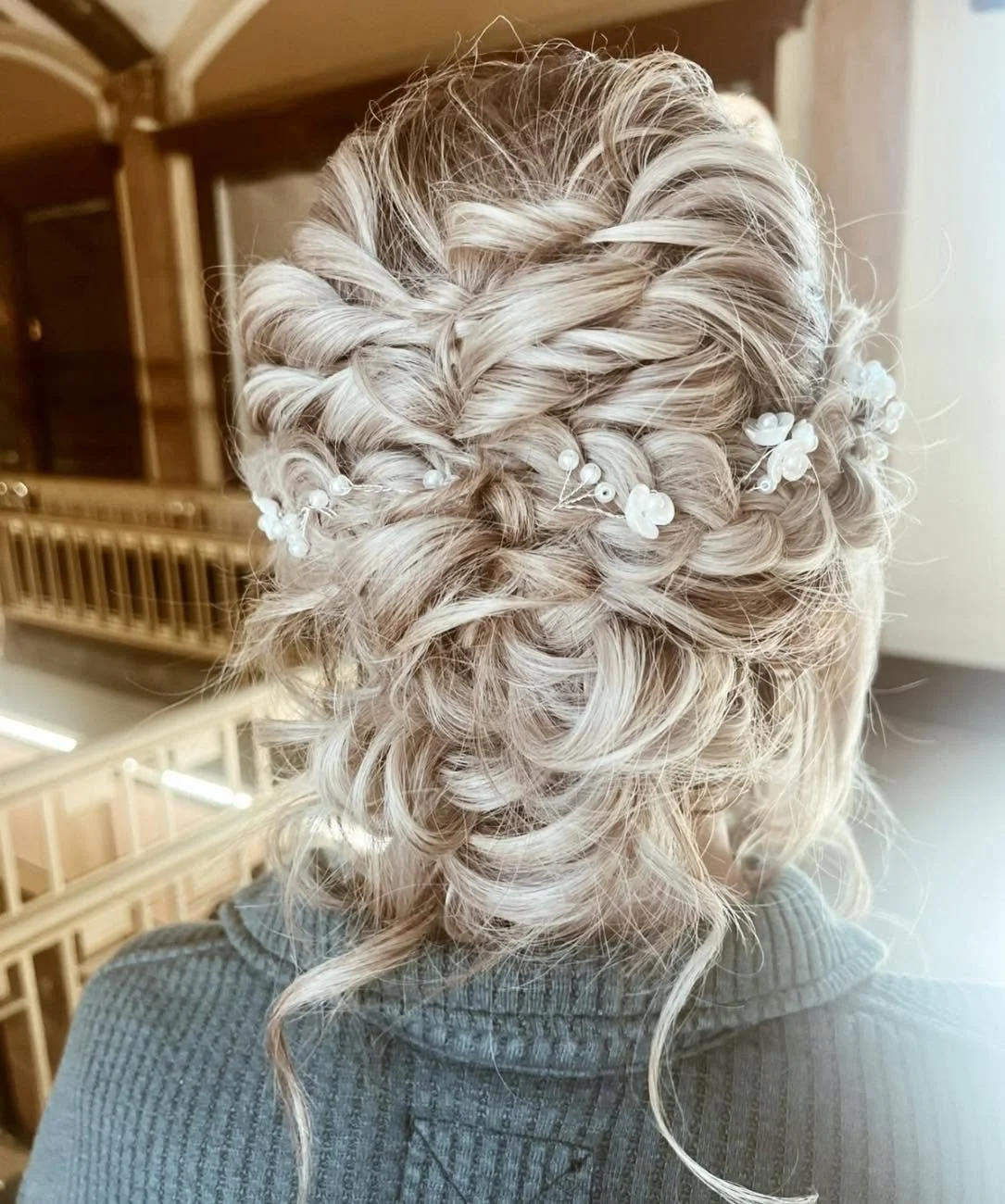 A woman with blonde, curly hair styled in an intricate updo with braid details and decorative white floral hair accessories.