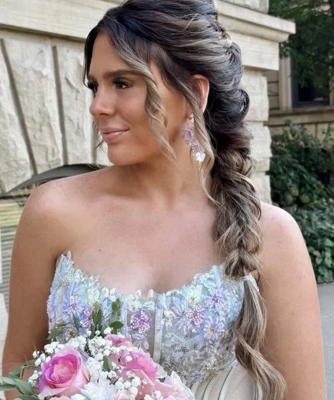 A woman in a white dress holding a bouquet of pink and white flowers, with long, styled hair and wearing earrings, standing outdoors in front of a stone building and greenery.