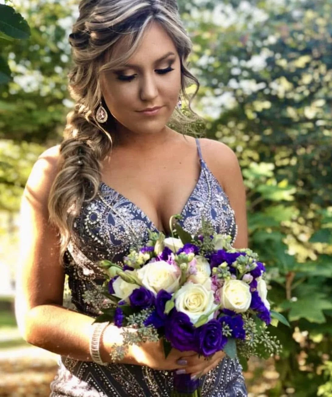 A woman with styled hair and earrings holds a bouquet of white and purple flowers outdoors, with trees and greenery in the background.