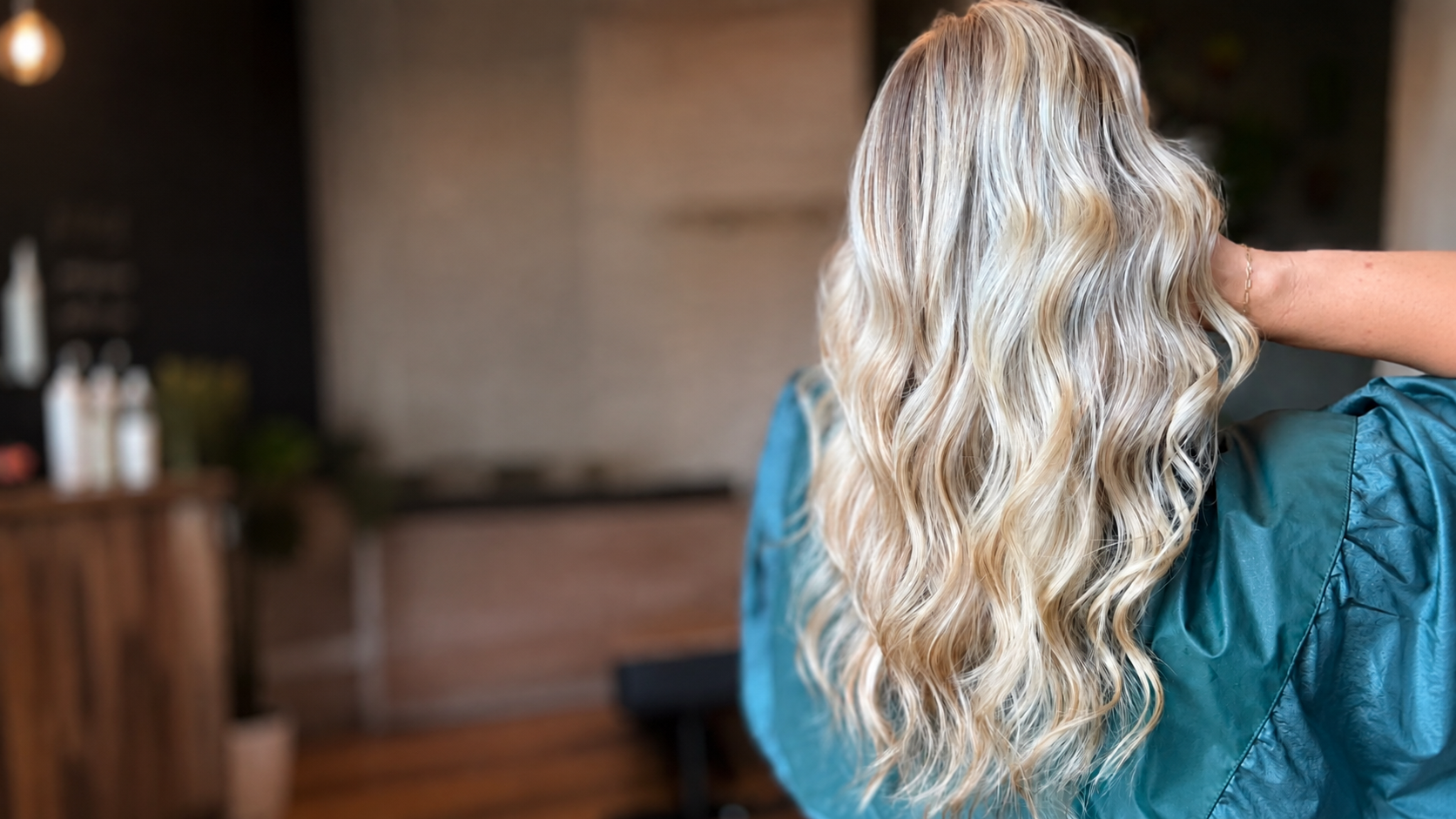 A woman with long, blonde, wavy hair in a salon, wearing a teal cape.