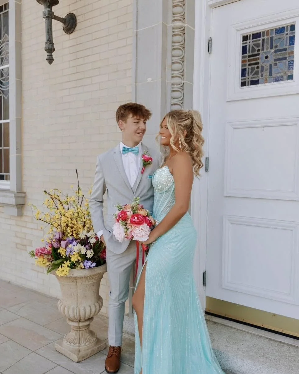 A young man and woman, dressed in formal attire, standing outside a building with a white door and brick wall, looking at each other. The man wears a light gray suit with a turquoise bow tie and boutonniere, while the woman wears a strapless, light b