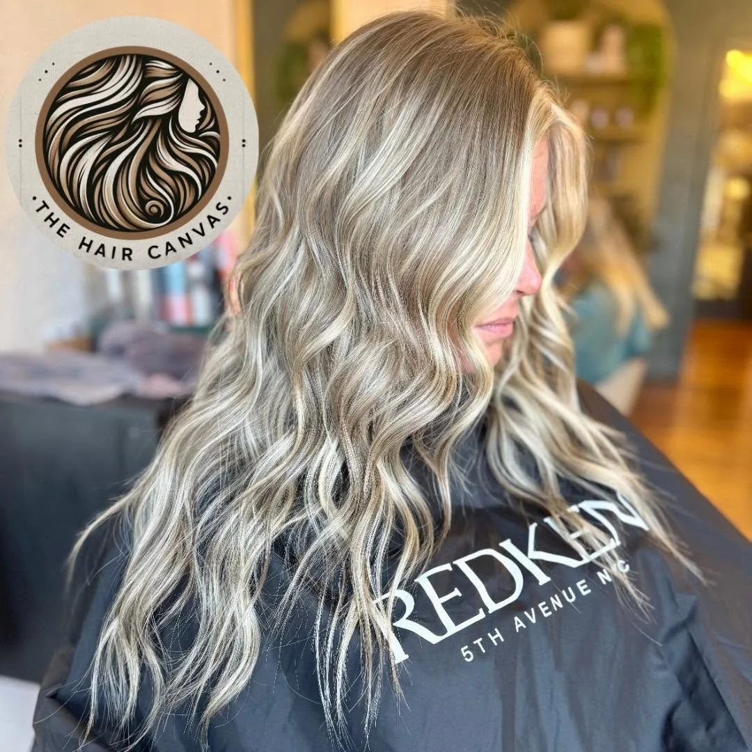 A woman with long, wavy blonde hair sitting at a salon, wearing a black cape with the words 'Redkissed 5th Avenue NYC' on it.