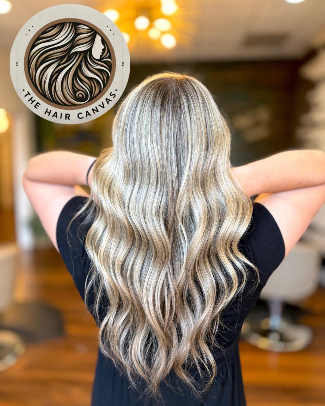 Back view of a woman with long, wavy blonde hair in a Salon. Logo in the top left corner says "The Hair Canvas" with an illustration of a woman's profile with hair.