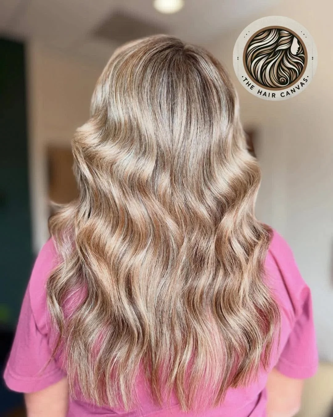Back view of a woman with long, wavy blonde hair in a salon. The hair has a shiny, smooth texture and a natural wave pattern. A logo for 'The Hair Canvas' is in the top right corner.