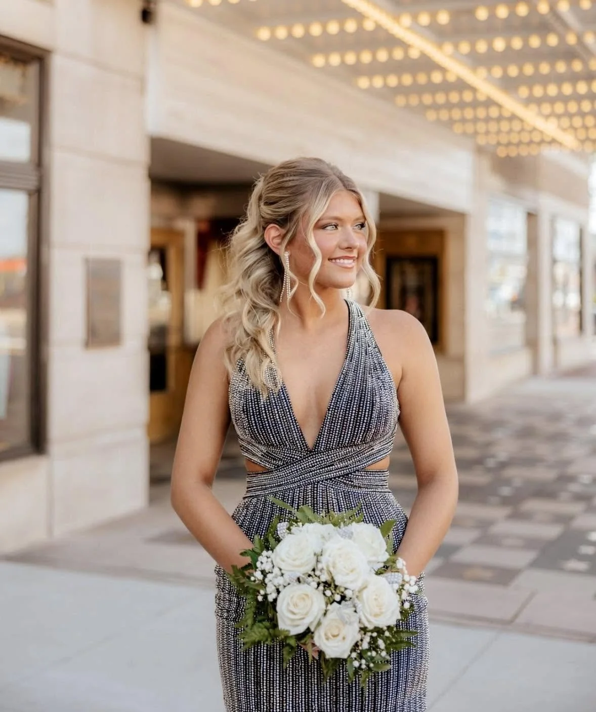 A young woman in a sparkly dress holding a bouquet of white roses standing outdoors at sunset with string lights overhead.