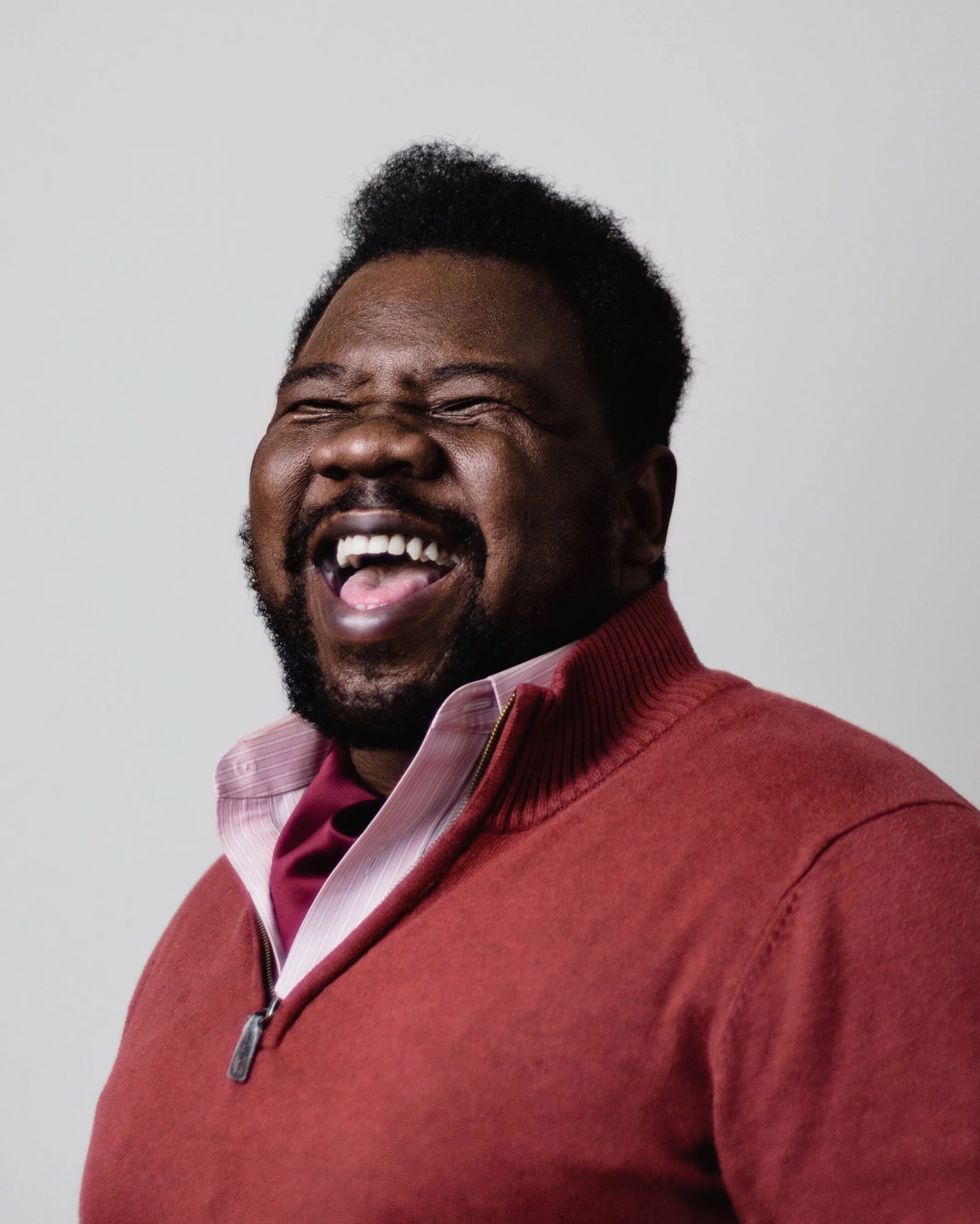 A close-up photo of a smiling man with dark skin, short curly hair, and a beard, wearing a red jacket over a pink striped shirt, against a plain light gray background.