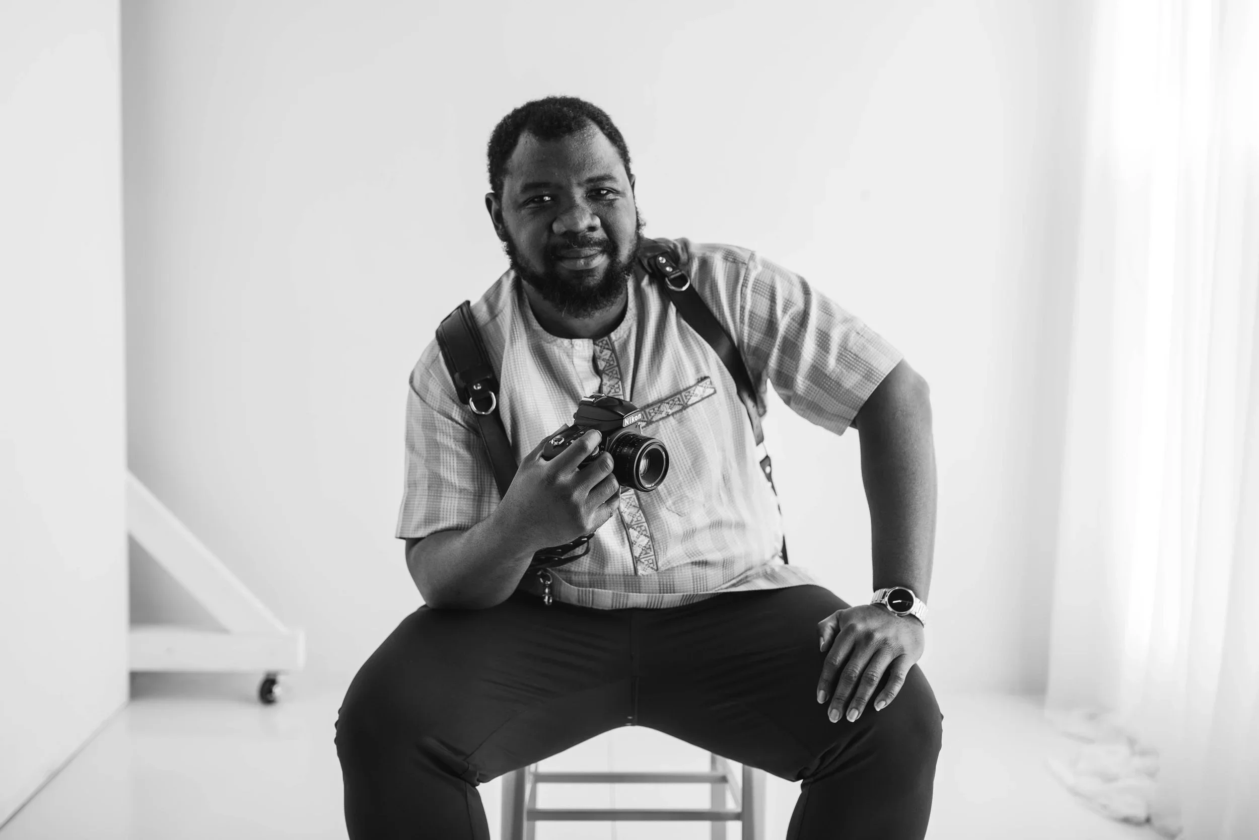 A man with a beard and mustache, wearing a light checkered shirt, sitting on a stool, holding a camera in his right hand, with a backpack strap over his shoulder, in a minimalist room with white walls.