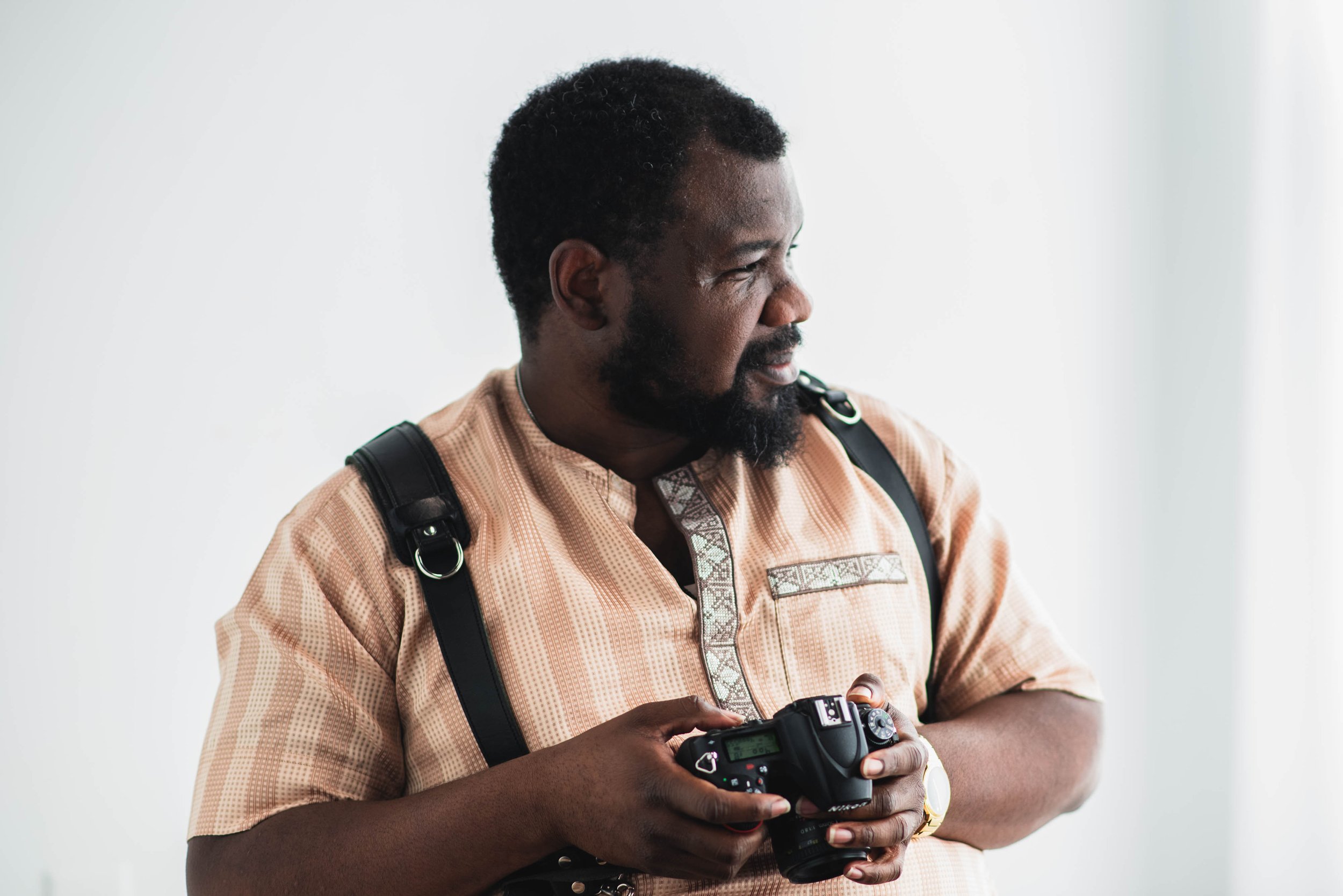A man of African descent with short curly hair and a beard is holding a camera, wearing a tan traditional shirt with intricate embroidery and black suspenders, standing against a plain white background.