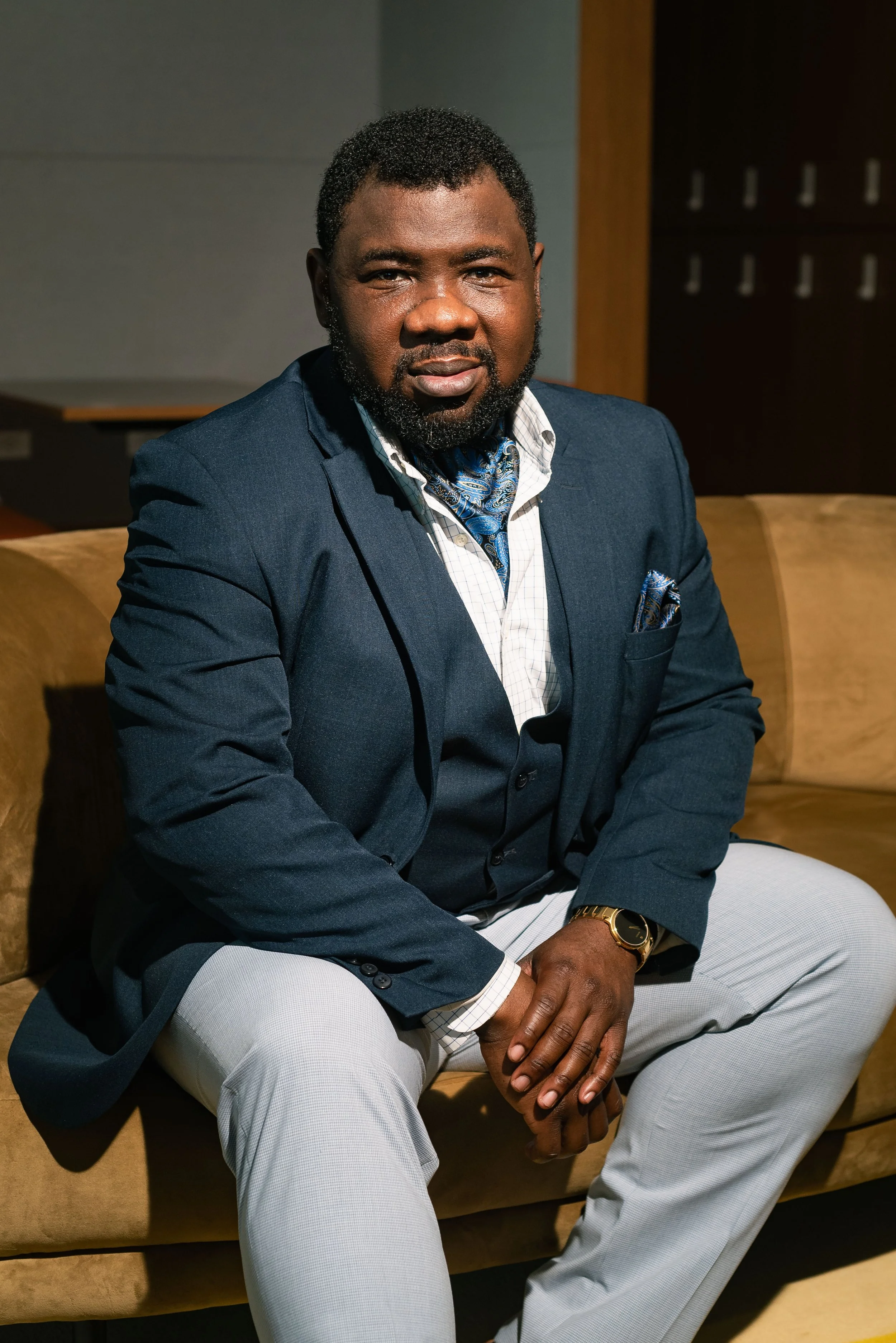 A well-dressed man with a beard and short curly hair sitting on a beige couch in an indoor setting, wearing a navy blazer, white shirt, patterned blue tie, grey trousers, and gold watch, with a confident expression.