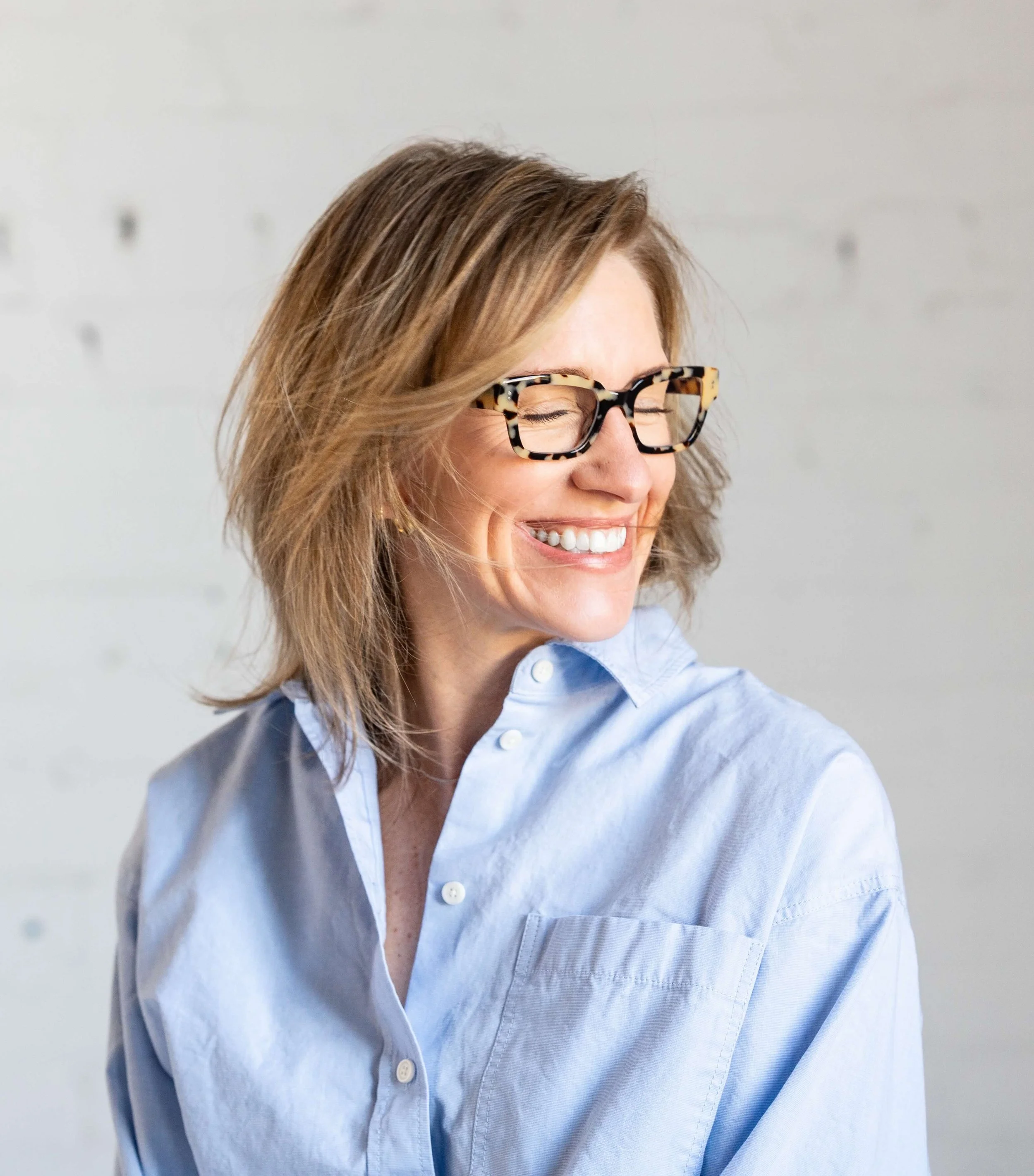 Close-up of a woman smiling with her eyes closed, wearing tortoiseshell glasses and a light blue button-up shirt, with light brown hair.