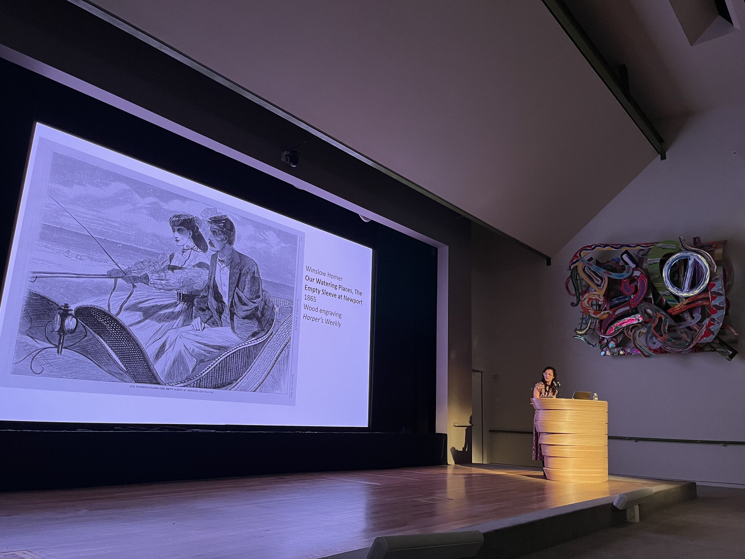 A person standing at a podium in front of a large framed illustration displayed on a projection screen. The illustration depicts two figures fishing: a woman and a man, with accompanying text describing it as Winslow Homer's wood engraving titled 'Our Watering Places, The Empty Sleeve at Newport, 1865.' The room has a high, angled ceiling, colorful abstract sculpture on the right, and a purpleish lighting ambiance.