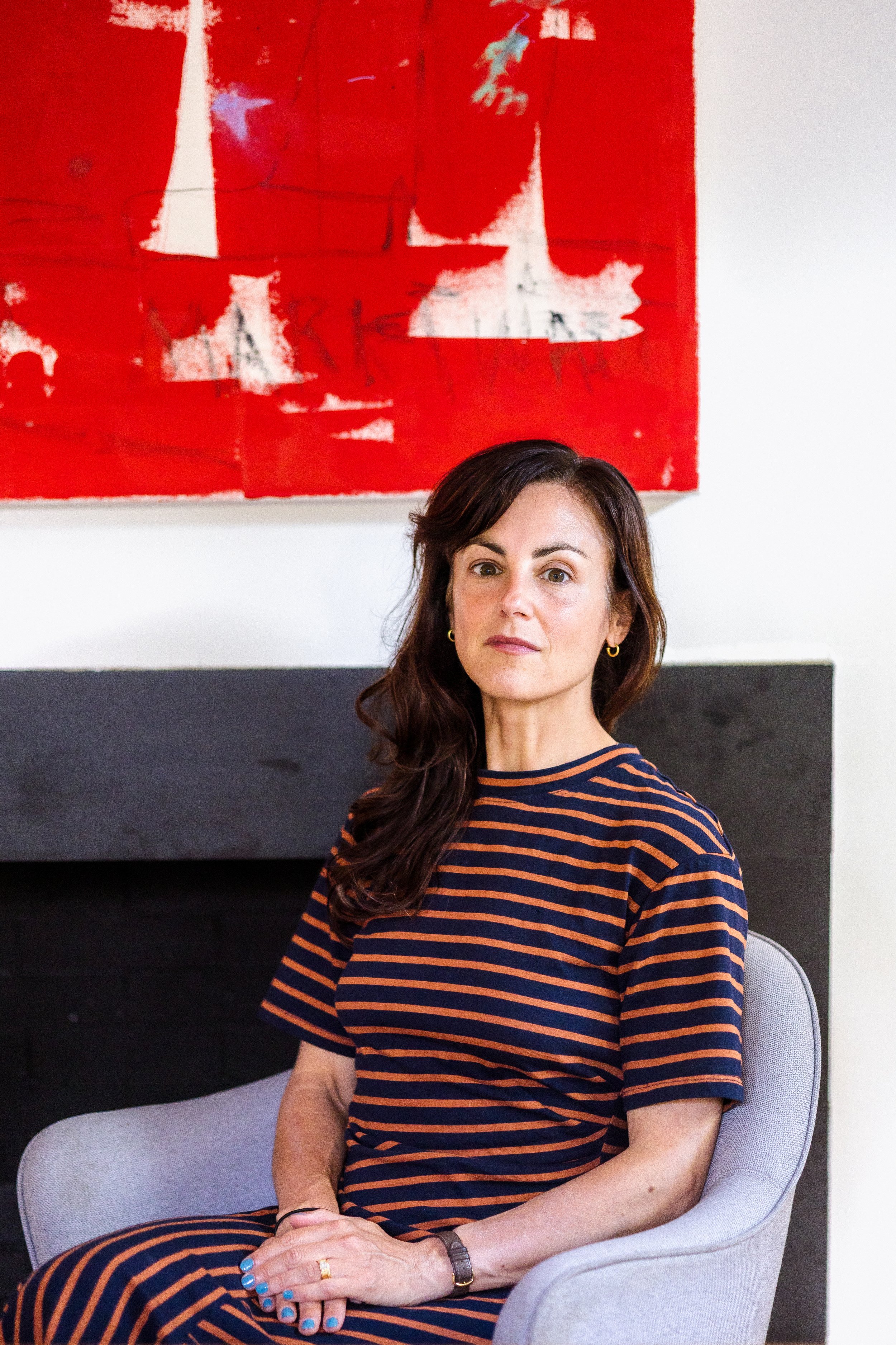A woman sitting on a light gray chair with black and orange striped dress, short brown hair, and blue nail polish, against a background with red, white, and black artwork on a white wall.