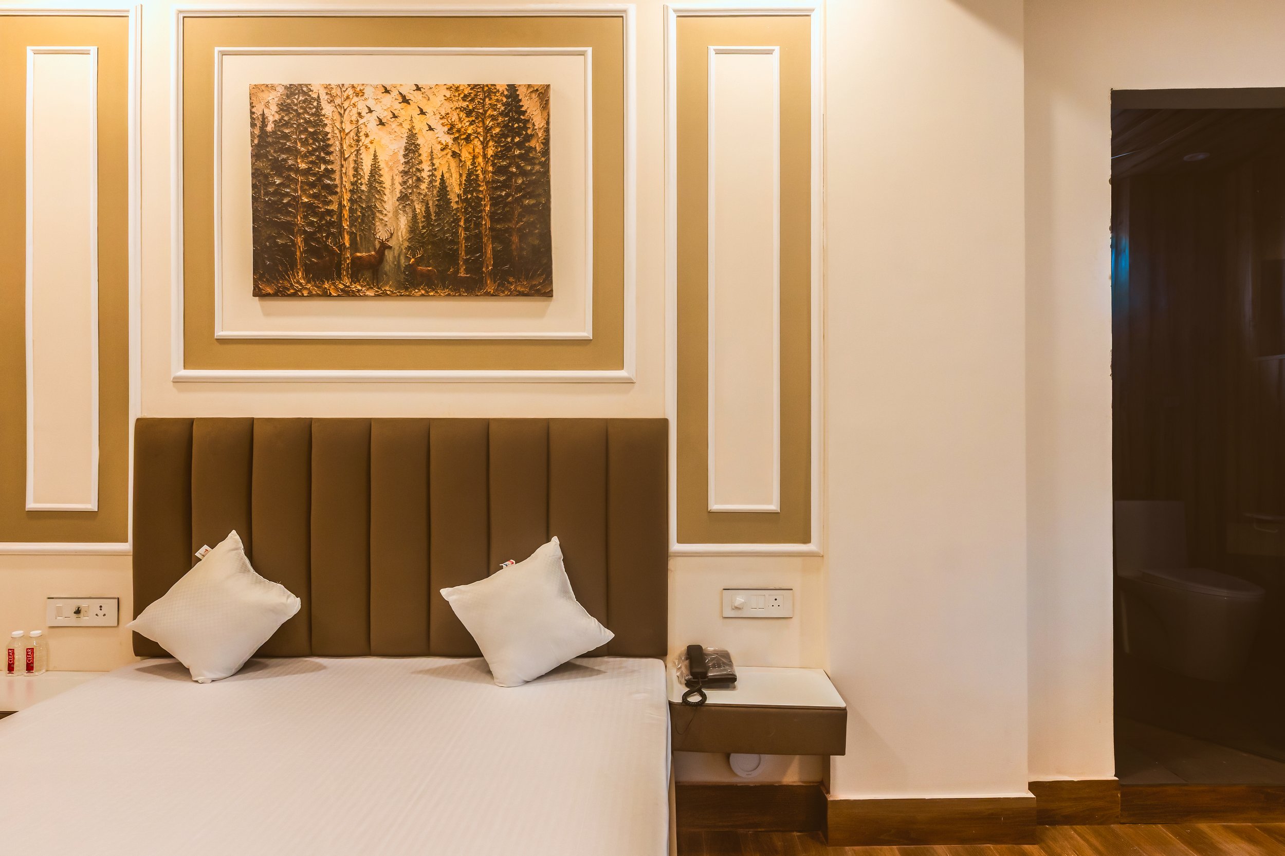 A hotel room with a bed, beige headboard, white bedding, and two pillows. A painting of a forest scene with deer is hung above the headboard. There are electrical outlets and a telephone on the bedside table.