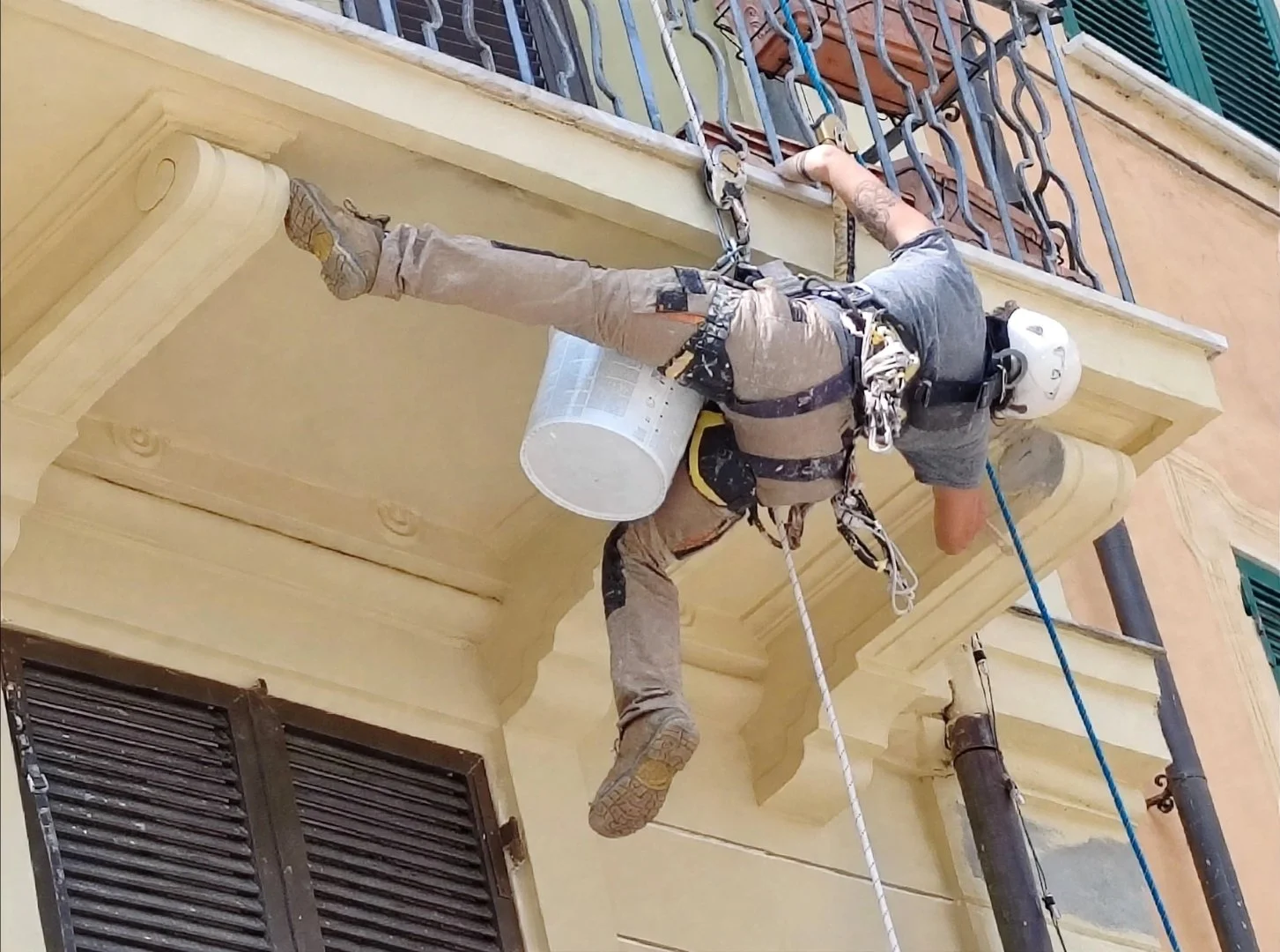 Lavoratore con imbracatura e casco mentre pulisce o lavora sotto un cornicione di un edificio su un balcone, utilizzando corde di sicurezza.