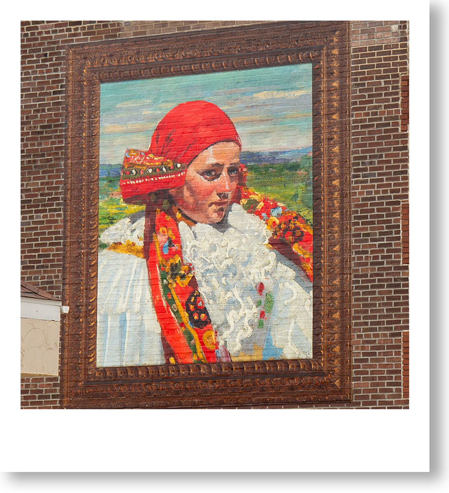A large mural on a brick building depicting a woman with a serious expression, wearing a red headscarf and a colorful traditional outfit, with a landscape background.