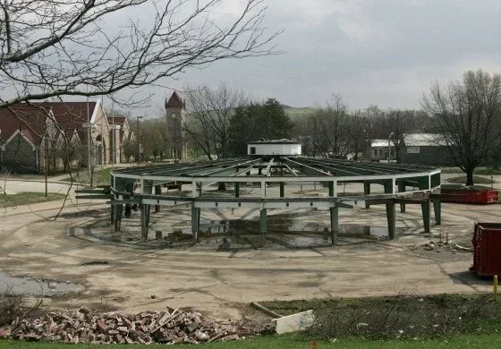 An empty, damaged roundabout in a suburban area with a partially dismantled railing, leafless trees, and overcast sky.