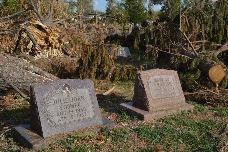 Two gravestones in a cemetery surrounded by fallen and damaged trees after a storm or natural disaster.