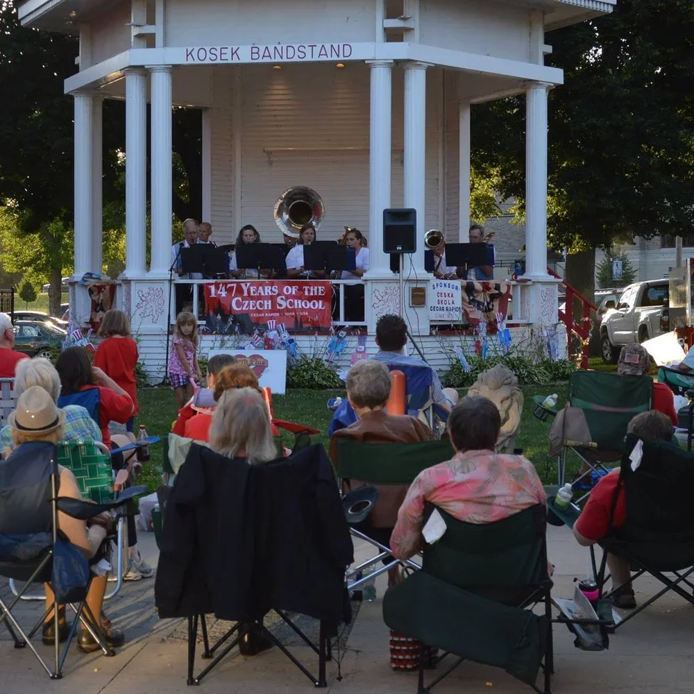 A band performing on a white pavilion stage at an outdoor event, with an audience sitting in portable chairs and some children standing nearby. The banner on the stage reads "147 Years of the Czech School." The pavilion has a sign that says "Kosek Ba
