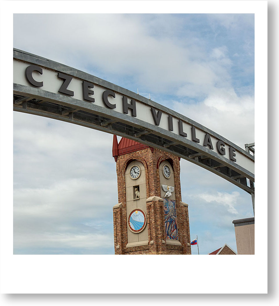 A clock tower with a red roof and brick walls, featuring weather-themed mosaics, is seen in the background. The foreground includes a partial sign reading "CZECH VILLAGE," arching overhead against a cloudy sky.