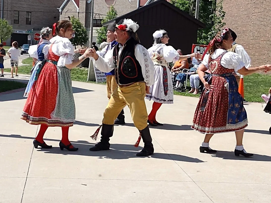 People dressed in traditional European folk costumes dancing and shaking hands outdoors during a cultural event.