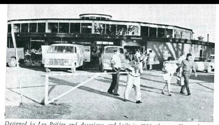 Black and white photo of a mid-century modern building with a curved glass facade, labeled 'ENTRANCE,' and a parking lot with several vintage cars. People are walking and socializing outside.