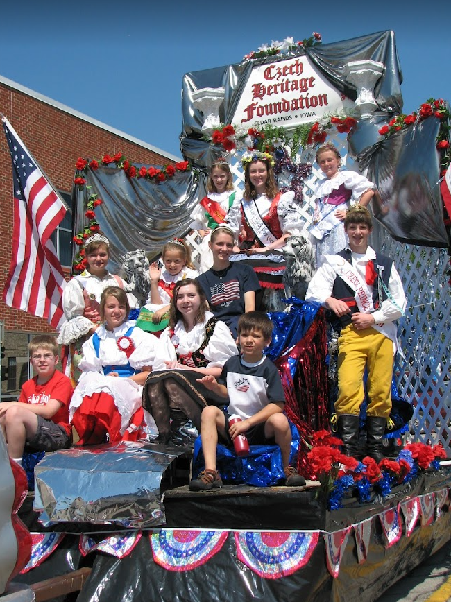 Children and a woman in traditional Czech attire on a parade float decorated with red, white, (American flag colors), and silver foil, under a sign that reads 'Czech Heritage Foundation, Cedar Rapids, Iowa', with American flags and onlookers nearby.