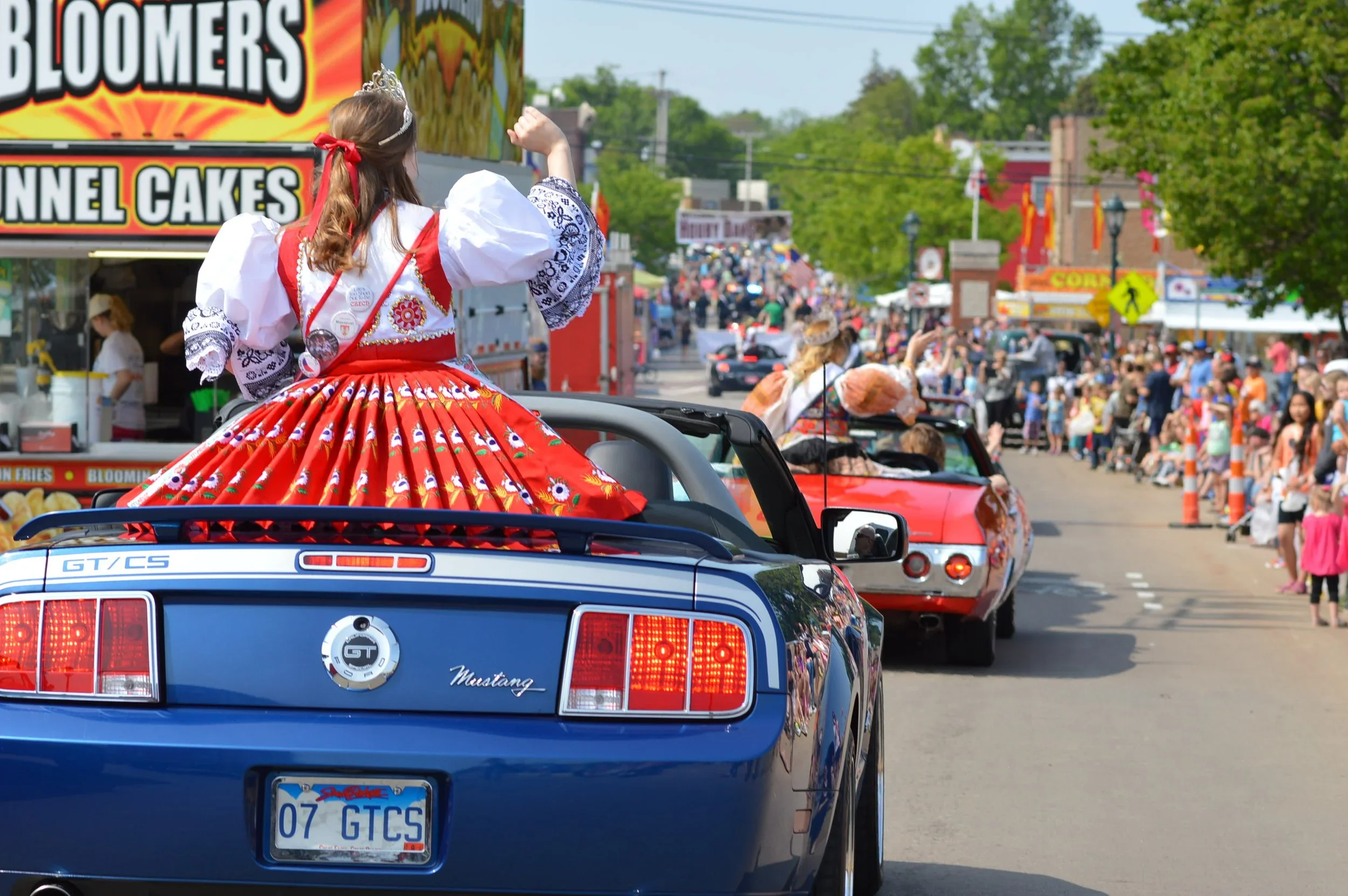 A parade with a woman dressed in a red, white, and black costume, wearing a crown and a sash, riding on the back of a blue Ford Mustang convertible. The street is lined with people, food stands, and colorful signs, with trees and buildings in the bac