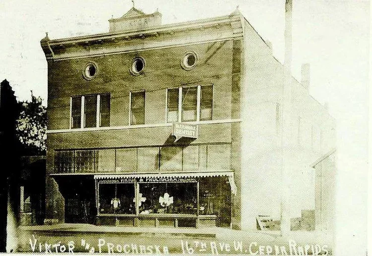 An old multi-story brick building with a storefront on the ground level, featuring large windows and a decorative awning. The building has three round windows on the upper floor and several rectangular windows on the upper levels.
