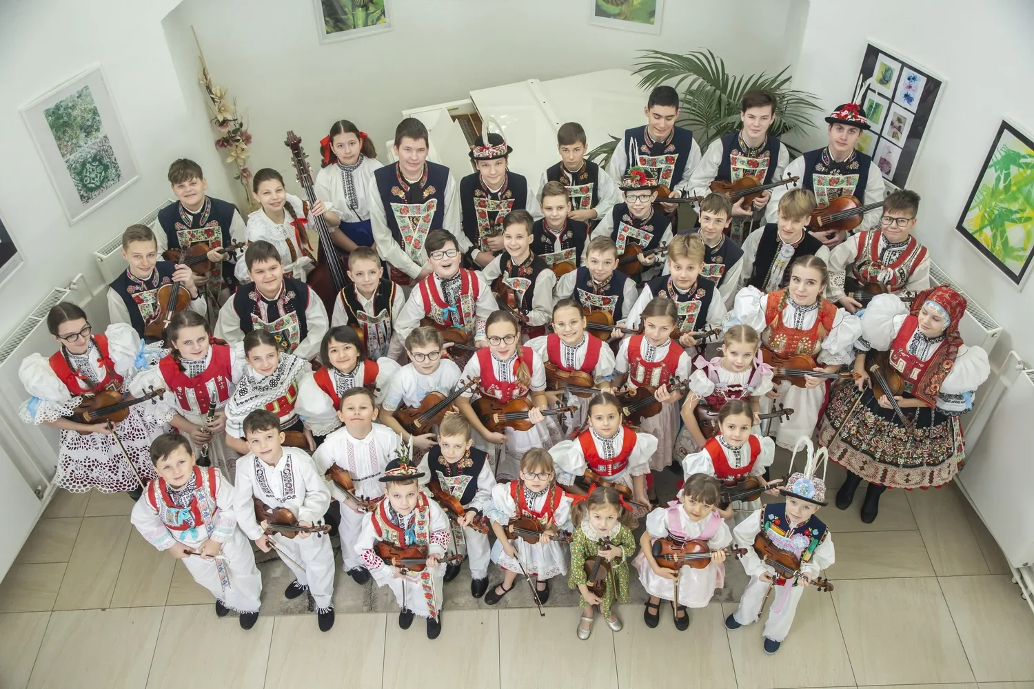 Children in traditional folk costumes holding violins in a room with white walls and colorful artwork.