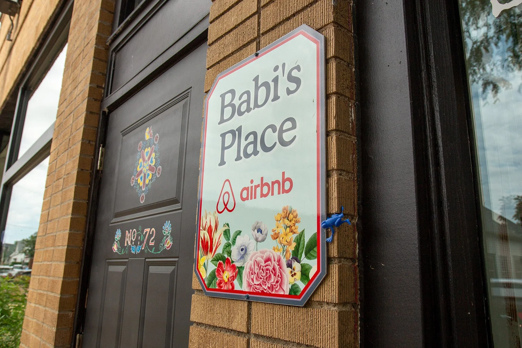 A sign hanging outside a building reads 'Babi's place' with the Airbnb logo, surrounded by colorful flowers, next to a black door and a brick wall.