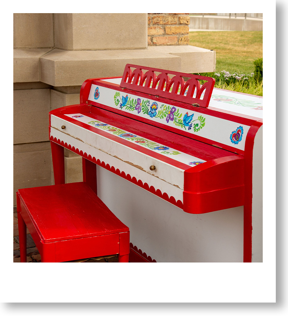 Colorful child-sized play piano with red, white, and floral patterns, and a matching red stool, outside near a stone wall and grass.