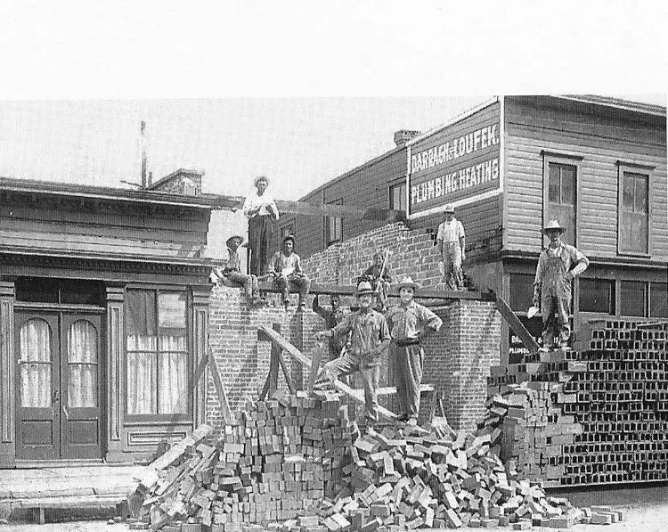 Black and white photo of construction workers standing and sitting on a partially built brick wall in front of wooden and brick buildings.