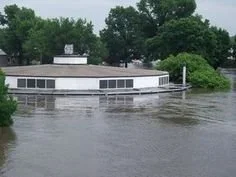 Flooded building surrounded by water and green trees in the background.