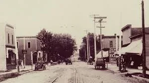 Historical black and white photo of a small town street with buildings on both sides, utility poles, a few trucks, and a tree in the background.