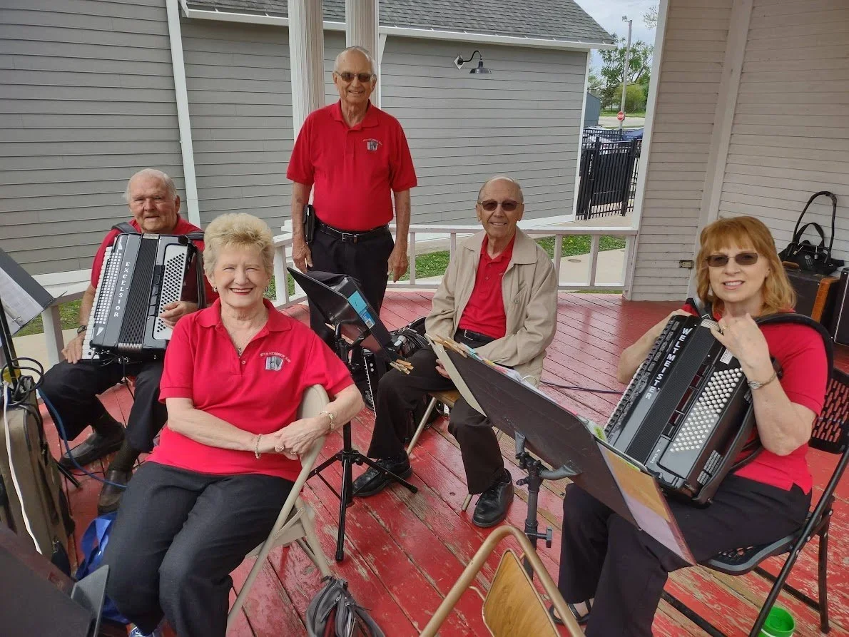 Five elderly people, three women and two men, are on a red wooden porch with musical instruments, including accordions and a music stand. They are smiling and appear ready to perform, dressed in red and neutral clothing.