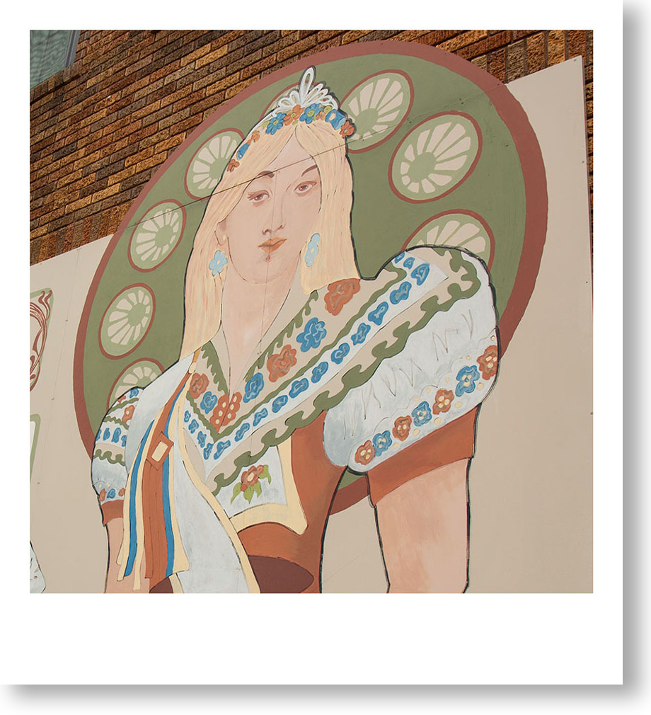 A painted mural of a woman with long blonde hair, wearing traditional folk clothing, with a decorative floral crown and a large circular design with lime slices behind her head.